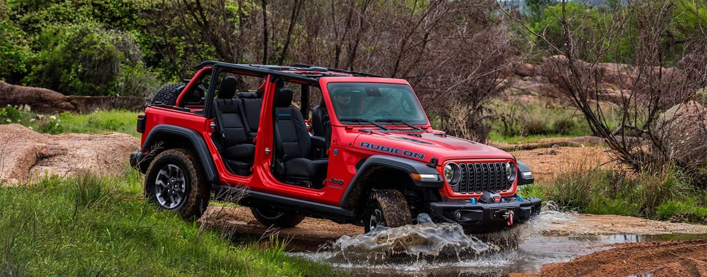 Side of a red 2026 Jeep Wrangler Rubicon driving over a shallow river.